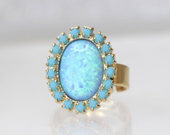 May include: A gold-toned ring featuring a large, oval-shaped, opalescent blue gemstone. The gemstone is surrounded by small, light blue, square-cut stones. The ring has an adjustable band.