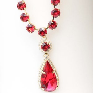 May include: A gold necklace with a teardrop-shaped pendant. The pendant is made of red and clear crystals. The necklace is adorned with red crystals.