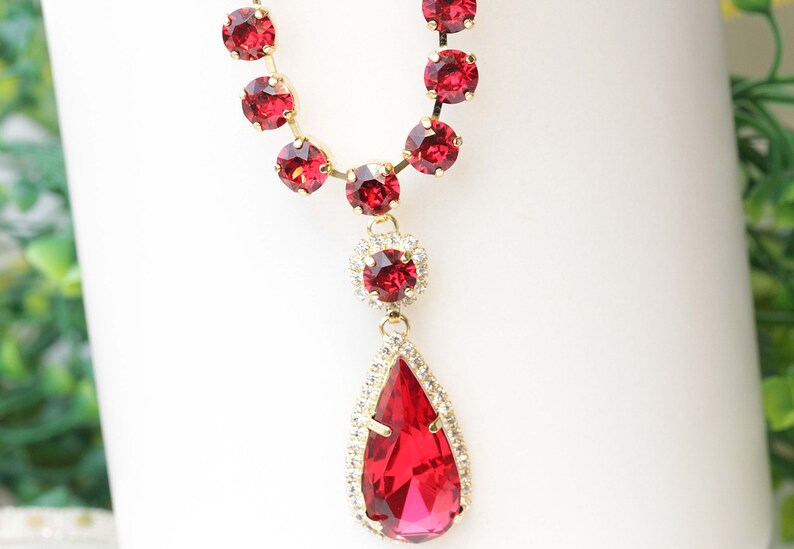May include: A gold necklace with a teardrop-shaped pendant. The pendant is made of red and clear crystals. The necklace is adorned with red crystals.