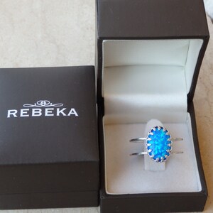 May include: A silver ring featuring a blue opal stone set within a decorative silver frame. The ring is presented in a black box with the inscription "REBEKA" on the lid.