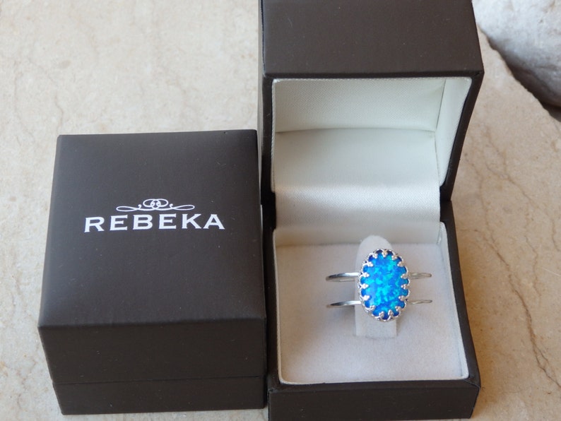 May include: A silver ring featuring a blue opal stone set within a decorative silver frame. The ring is presented in a black box with the inscription "REBEKA" on the lid.