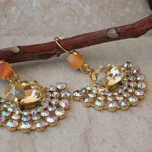 May include: A pair of gold earrings with a large clear crystal and a smaller peach-colored crystal. The earrings are surrounded by smaller clear crystals.