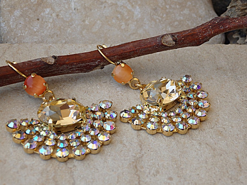 May include: A pair of gold earrings with a large clear crystal and a smaller peach-colored crystal. The earrings are surrounded by smaller clear crystals.