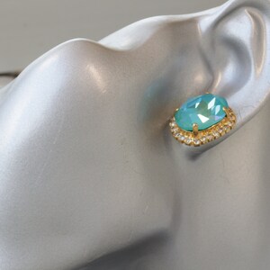 Puede incluir: Un par de pendientes de oro con una piedra ovalada de color azul claro rodeada por un halo de piedras preciosas m&aacute;s peque&ntilde;as y transparentes.