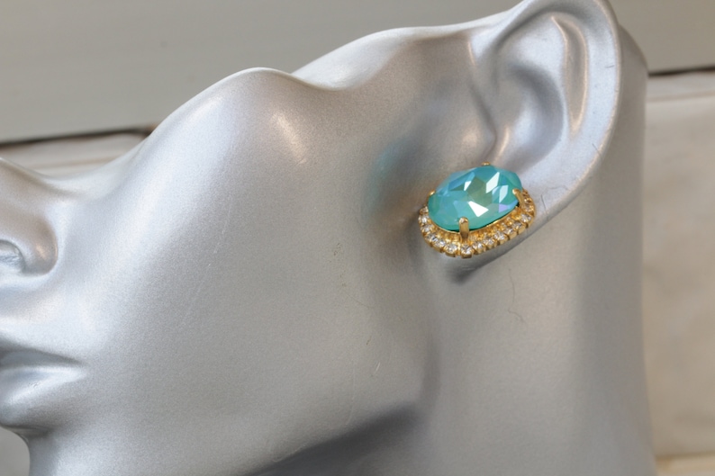 Puede incluir: Un par de pendientes de oro con una piedra ovalada de color azul claro rodeada por un halo de piedras preciosas m&aacute;s peque&ntilde;as y transparentes.