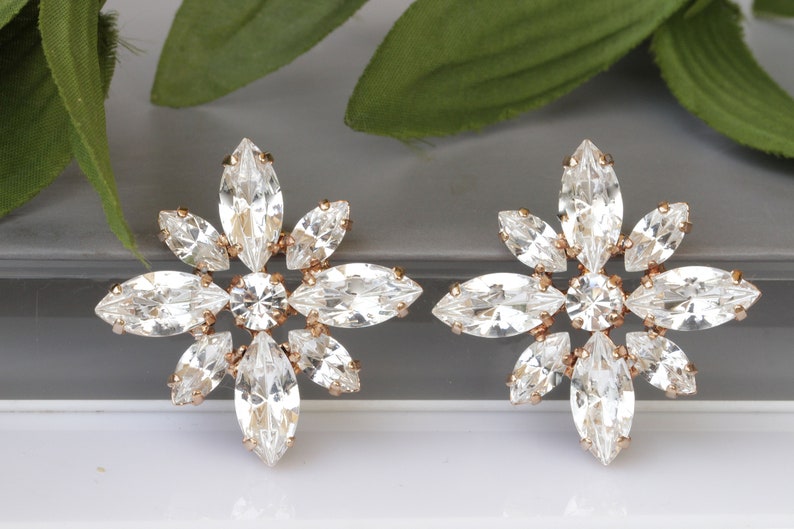 May include: A pair of rose gold earrings with a floral design. Each earring features a cluster of clear marquise-shaped crystals arranged in a starburst pattern.