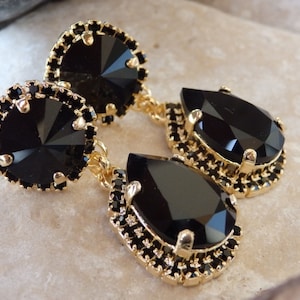 May include: A pair of gold tone dangle earrings with black teardrop shaped crystals. The crystals are surrounded by a gold tone setting with smaller black crystals.