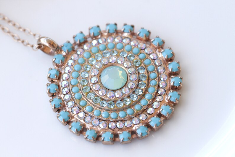 May include: A gold necklace with a large, round pendant featuring a turquoise and clear crystal design. The pendant is made up of concentric circles of turquoise and clear crystals, with a larger, clear crystal in the center.