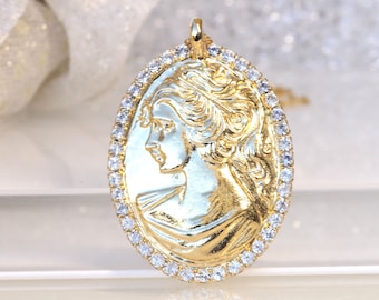 May include: A gold oval pendant featuring a detailed profile of a woman's face. The pendant is bordered by a row of clear cubic zirconia stones.