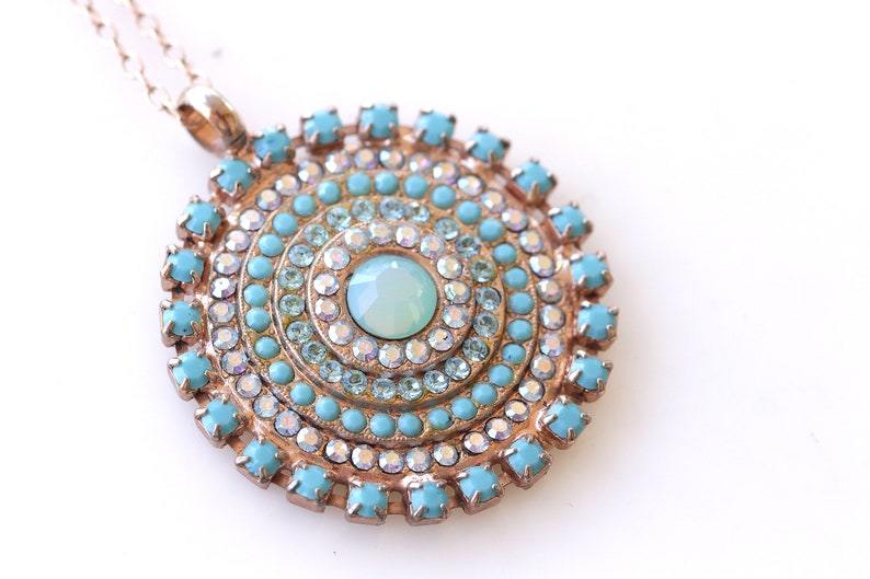 May include: A gold necklace with a round pendant featuring a turquoise and clear crystal design. The pendant is made up of concentric circles of turquoise and clear crystals.