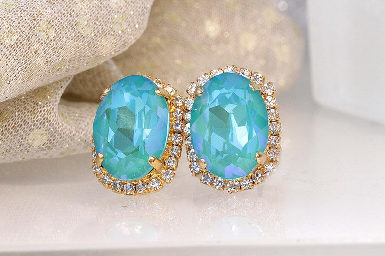 Puede incluir: Un par de pendientes ovalados con una piedra preciosa azul claro rodeada por un halo de brillantes transparentes. Los pendientes tienen un engaste de oro.