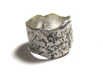 May include: A silver ring with a textured, hammered finish. The ring is wide and has a unique, organic shape.