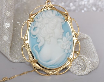 Puede incluir: Un colgante de camafeo en tono dorado con un camafeo azul y blanco de un perfil de mujer. El camafeo está engastado en un marco en tono dorado con un diseño ornamentado.
