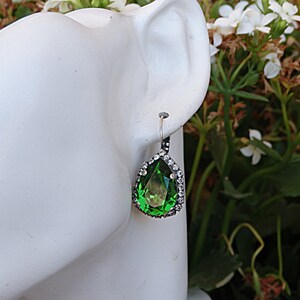 May include: A pair of dangle earrings with a pear-shaped green gemstone surrounded by a halo of clear rhinestones. The earrings are silver-toned and have a hook closure.