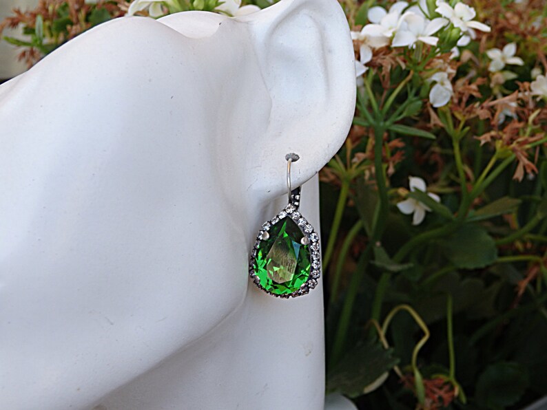 May include: A pair of dangle earrings with a pear-shaped green gemstone surrounded by a halo of clear rhinestones. The earrings are silver-toned and have a hook closure.