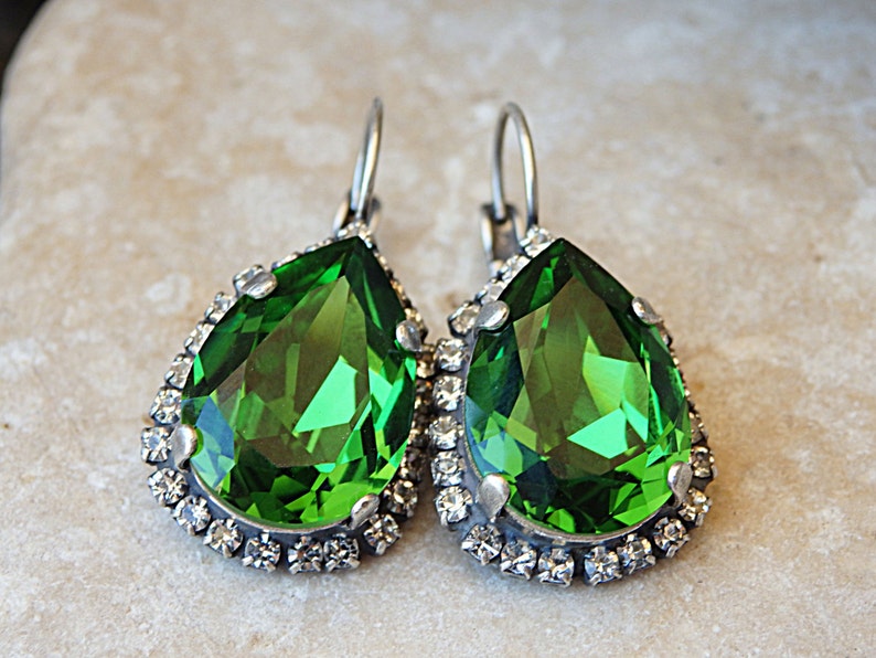 May include: A pair of dangle earrings with a large pear-shaped green gemstone surrounded by a halo of smaller clear rhinestones.