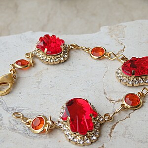 May include: Gold bracelet featuring three red hamsa hand charms with orange crystal accents.