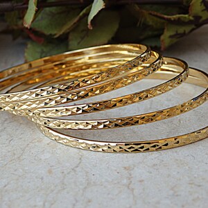 Può includere: Set di cinque braccialetti dorati con un motivo a forma di diamante. I braccialetti sono impilati uno sopra l'altro.