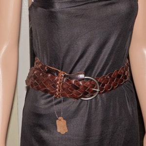 May include: A brown leather braided belt with a silver buckle. The belt is worn over a black dress.