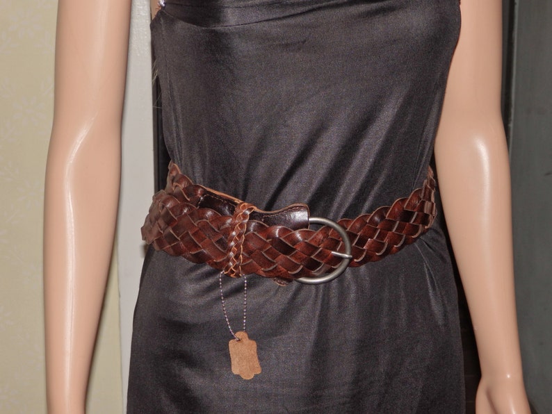 May include: A brown leather braided belt with a silver buckle. The belt is worn over a black dress.
