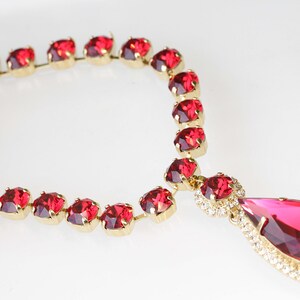 May include: A red rhinestone necklace with a teardrop pendant. The necklace is made of gold-toned metal and features a row of red rhinestones.
