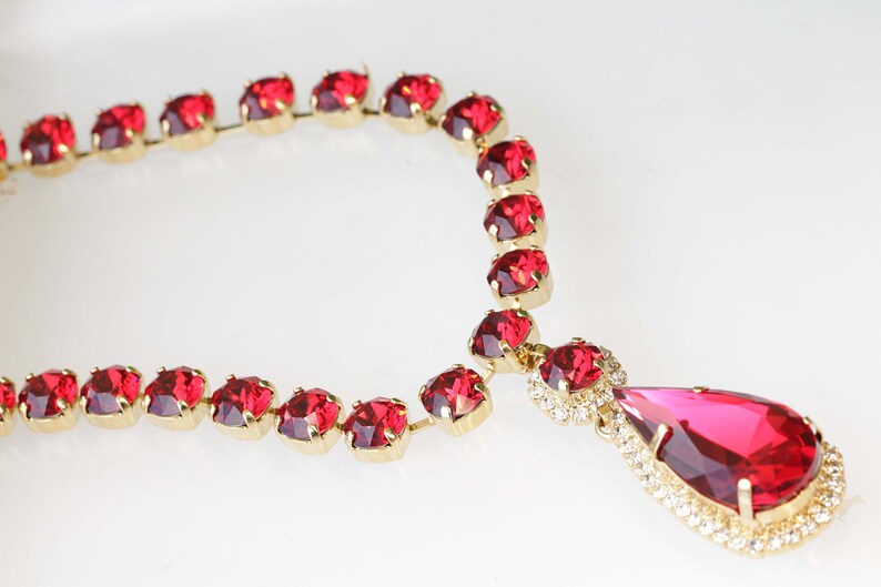 May include: A red rhinestone necklace with a teardrop pendant. The necklace is made of gold-toned metal and features a row of red rhinestones.
