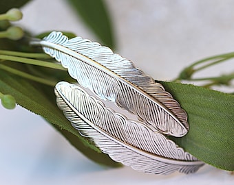 May include: Silver bracelet with a double feather design. The feathers are detailed with a textured pattern.