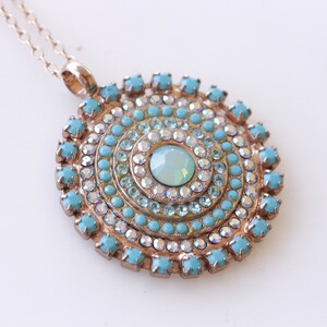 May include: A gold necklace with a round pendant featuring a turquoise and clear crystal design. The pendant is made up of concentric circles of crystals.