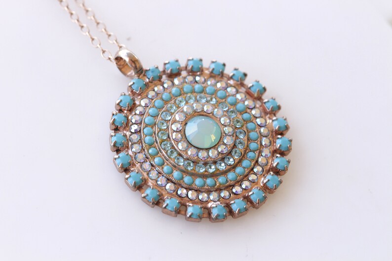 May include: A gold necklace with a round pendant featuring a turquoise and clear crystal design. The pendant is made up of concentric circles of crystals.