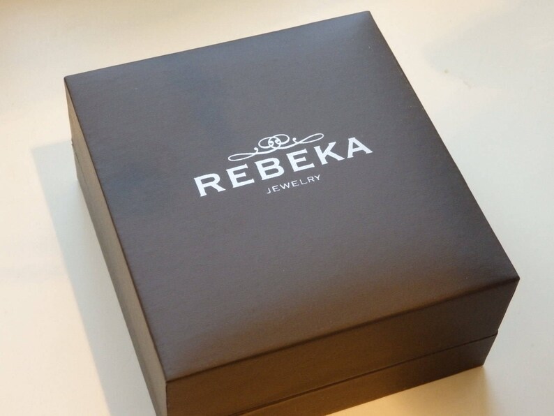 Puede incluir: Una caja rectangular de color gris oscuro con el texto "REBEKA JEWELRY" en letras blancas. La caja tiene un dise&ntilde;o decorativo con un c&iacute;rculo doble estilizado.