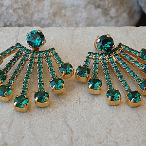 May include: A pair of gold earrings with emerald green crystal accents. The earrings feature a large round crystal in the center and smaller crystals arranged in a fan shape.