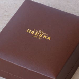 May include: A brown rectangular box with the text "REBEKA JEWELRY" in gold lettering.