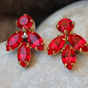 Puede incluir: Un par de pendientes de oro con detalles de cristal rojo. Los pendientes presentan un grupo de cinco cristales rojos, con un cristal redondo más grande en la parte superior.