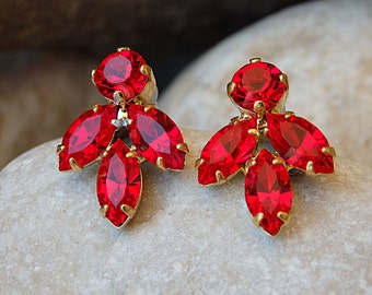 May include: A pair of gold earrings with red crystal accents. The earrings feature a cluster of five red crystals, with a larger round crystal at the top.