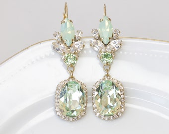 Może przedstawiać: Para kolczyków wiszących z zielonym i białym wzorem z kryształów. Kolczyki mają duży owalny zielony kryształ otoczony mniejszymi przezroczystymi kryształami oraz mniejszy zielony kryształ z przezroczystymi kryształami nad nim.