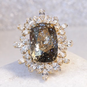 May include: A large, faceted, dark brown gemstone ring with a gold setting. The ring is surrounded by a halo of smaller, clear gemstones.