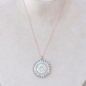 May include: A rose gold chain necklace with a round pendant featuring a concentric circle design of light blue and white rhinestones.