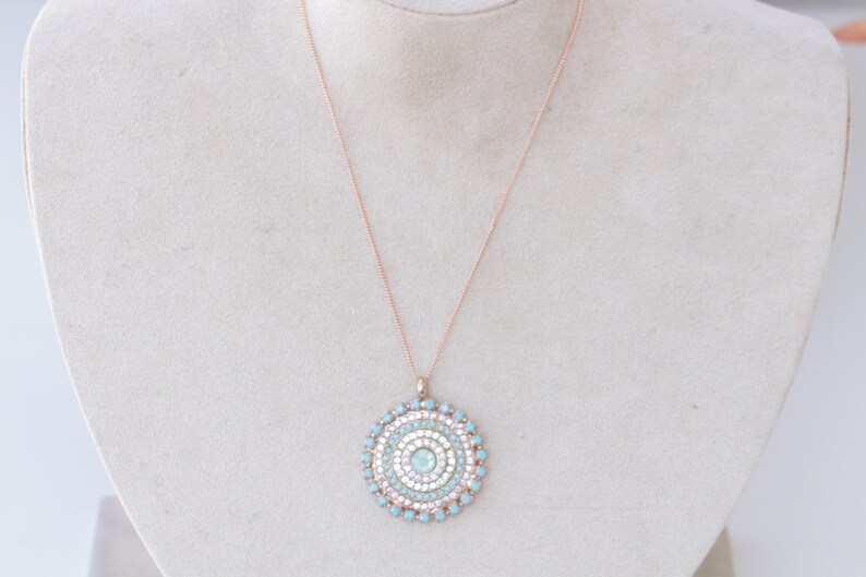 May include: A rose gold chain necklace with a round pendant featuring a concentric circle design of light blue and white rhinestones.