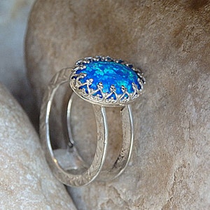 May include: A silver ring featuring a blue opal gemstone set within a filigree design. The ring is displayed on a textured surface.