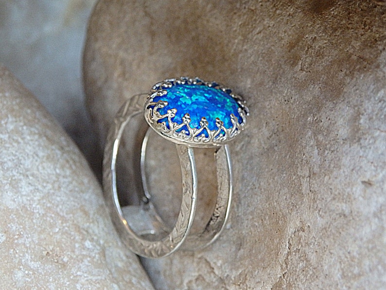 May include: A silver ring featuring a blue opal gemstone set within a filigree design. The ring is displayed on a textured surface.