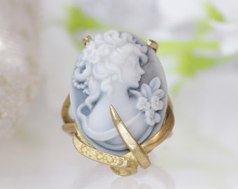 Può includere: Un anello d'oro con un cammeo che raffigura il profilo di una donna in bianco e grigio. Il cammeo è incastonato in una cornice d'oro con un motivo floreale.