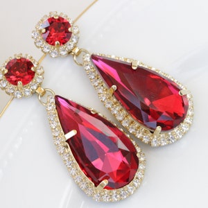 May include: A pair of dangle earrings with large teardrop-shaped red crystals surrounded by a halo of smaller clear crystals. The earrings are gold-toned.