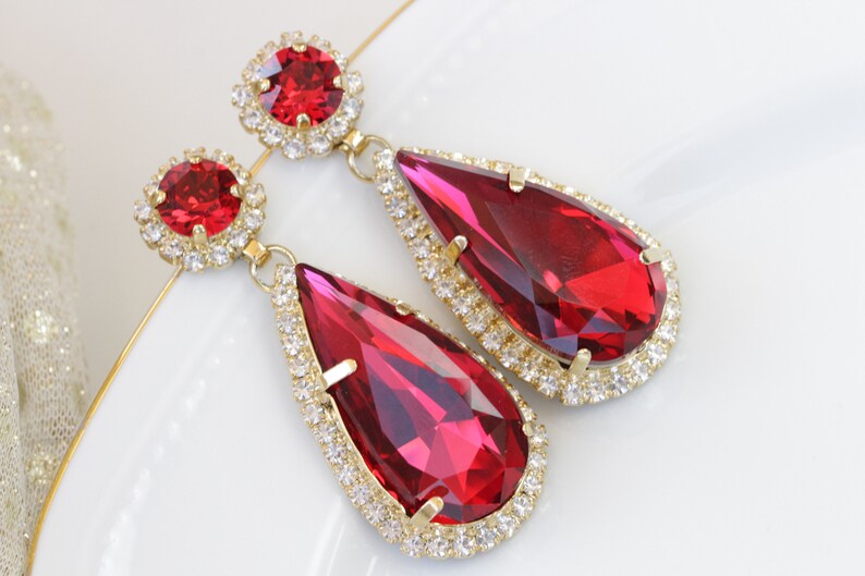 May include: A pair of dangle earrings with large teardrop-shaped red crystals surrounded by a halo of smaller clear crystals. The earrings are gold-toned.