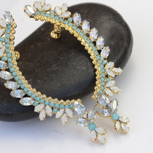 May include: A gold ear cuff with white and light blue rhinestones. The cuff is shaped like a crescent moon and has a dangling piece with more rhinestones.