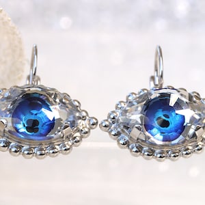 May include: A pair of silver earrings featuring clear crystal stones with a blue evil eye design.