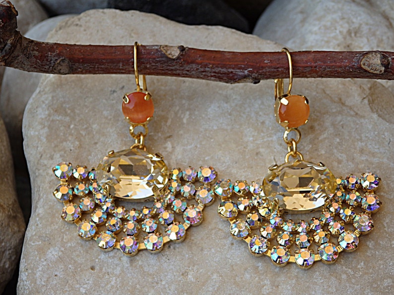 May include: A pair of gold earrings with a large clear crystal and a smaller orange stone. The earrings are decorated with a fringe of smaller crystals.