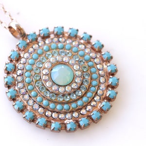 May include: A gold necklace with a round pendant featuring a turquoise and clear crystal design. The pendant is made up of concentric circles of turquoise and clear crystals.
