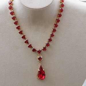 May include: A red rhinestone necklace with a teardrop pendant. The necklace is made of gold-tone metal and features a row of round red rhinestones.