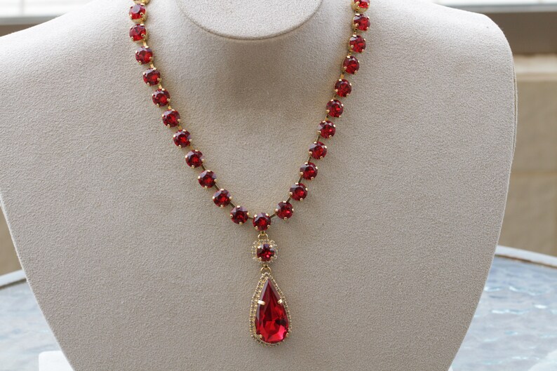 May include: A red rhinestone necklace with a teardrop pendant. The necklace is made of gold-tone metal and features a row of round red rhinestones.