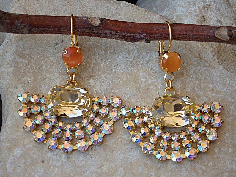 May include: A pair of gold tone earrings with a large clear crystal and a smaller peach colored stone. The earrings are dangling and have a fan-shaped design with smaller crystals.